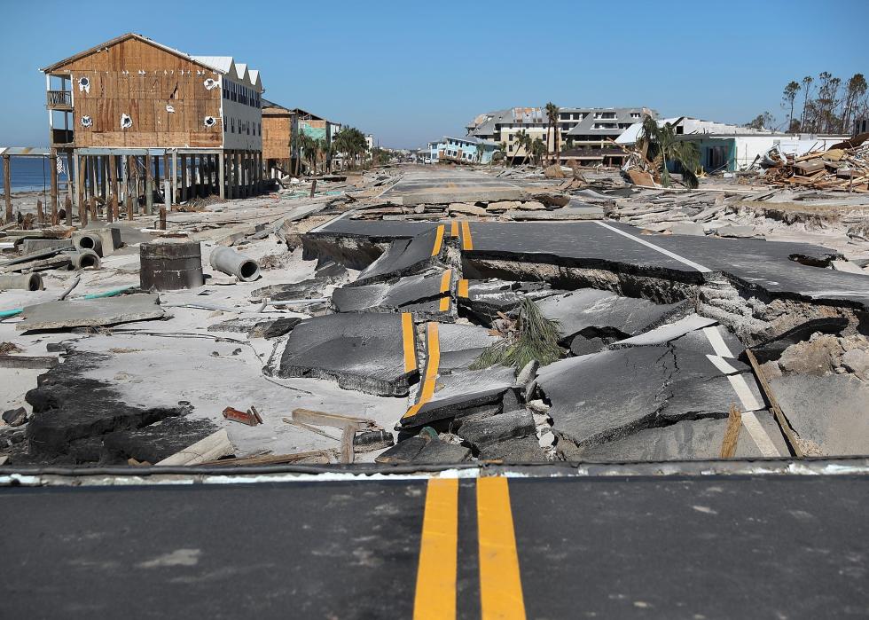 A blue sky in the background and in the forefront, a black asphalt road with freshly painted bright yellow lines in pieces near the beach and surrounded by homes that have also been badly damaged by a storm.