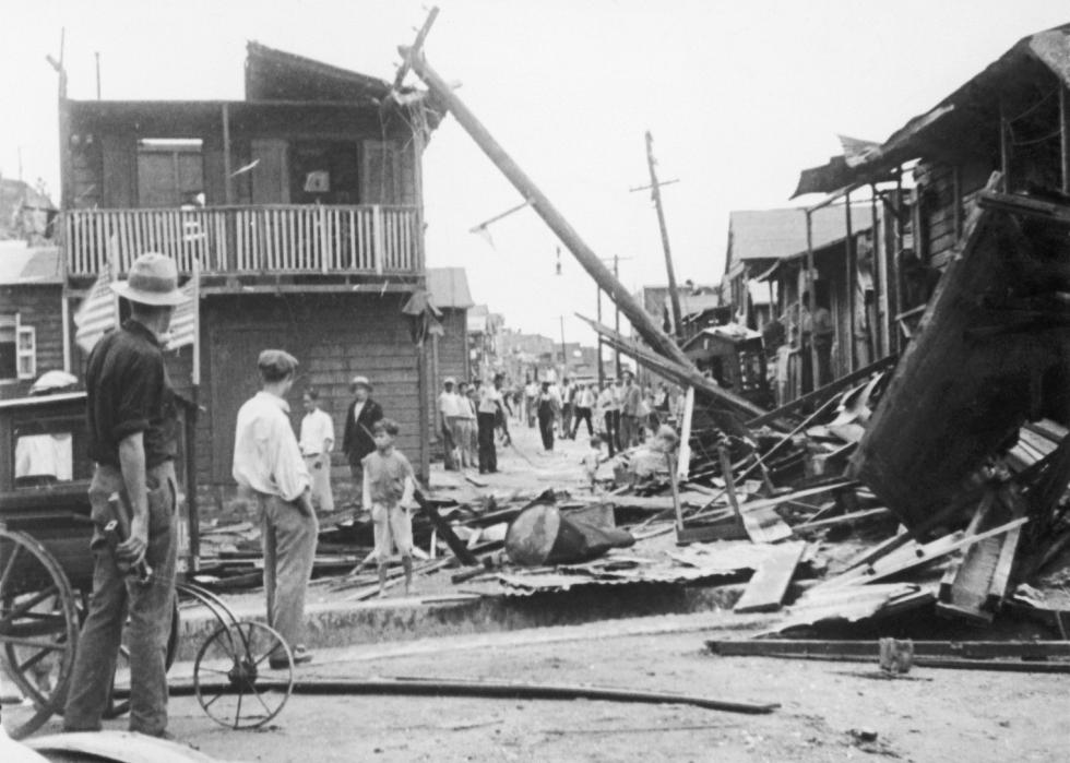 People standing along a street of destructed two-storey buildings. 