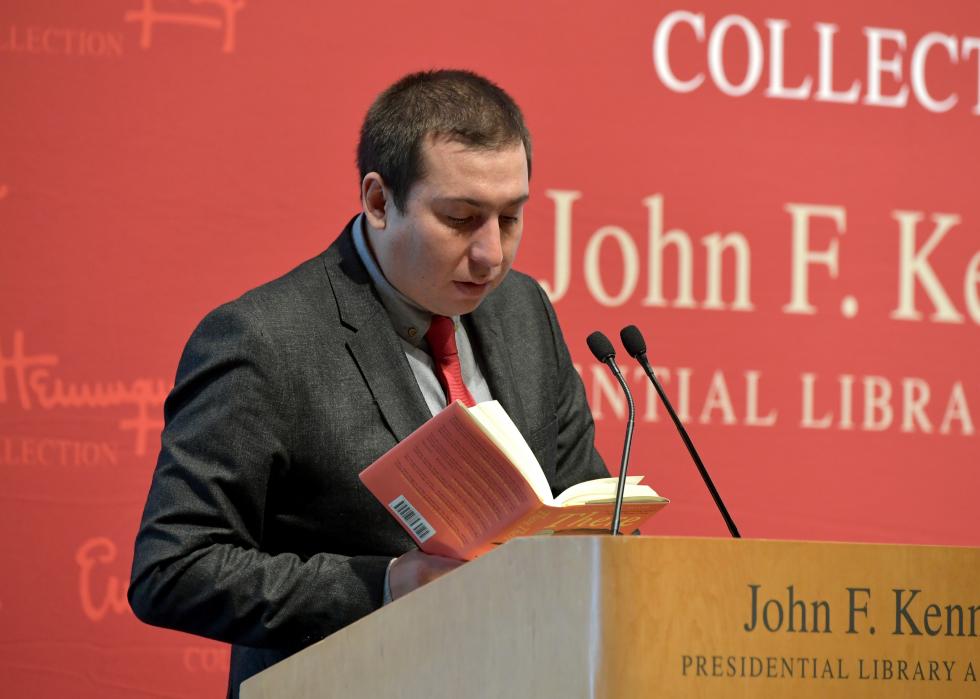 Tommy Orange, standing behind a podium at the John F. Kennedy Presidential Library and Museum, reads aloud from his book into a mic.