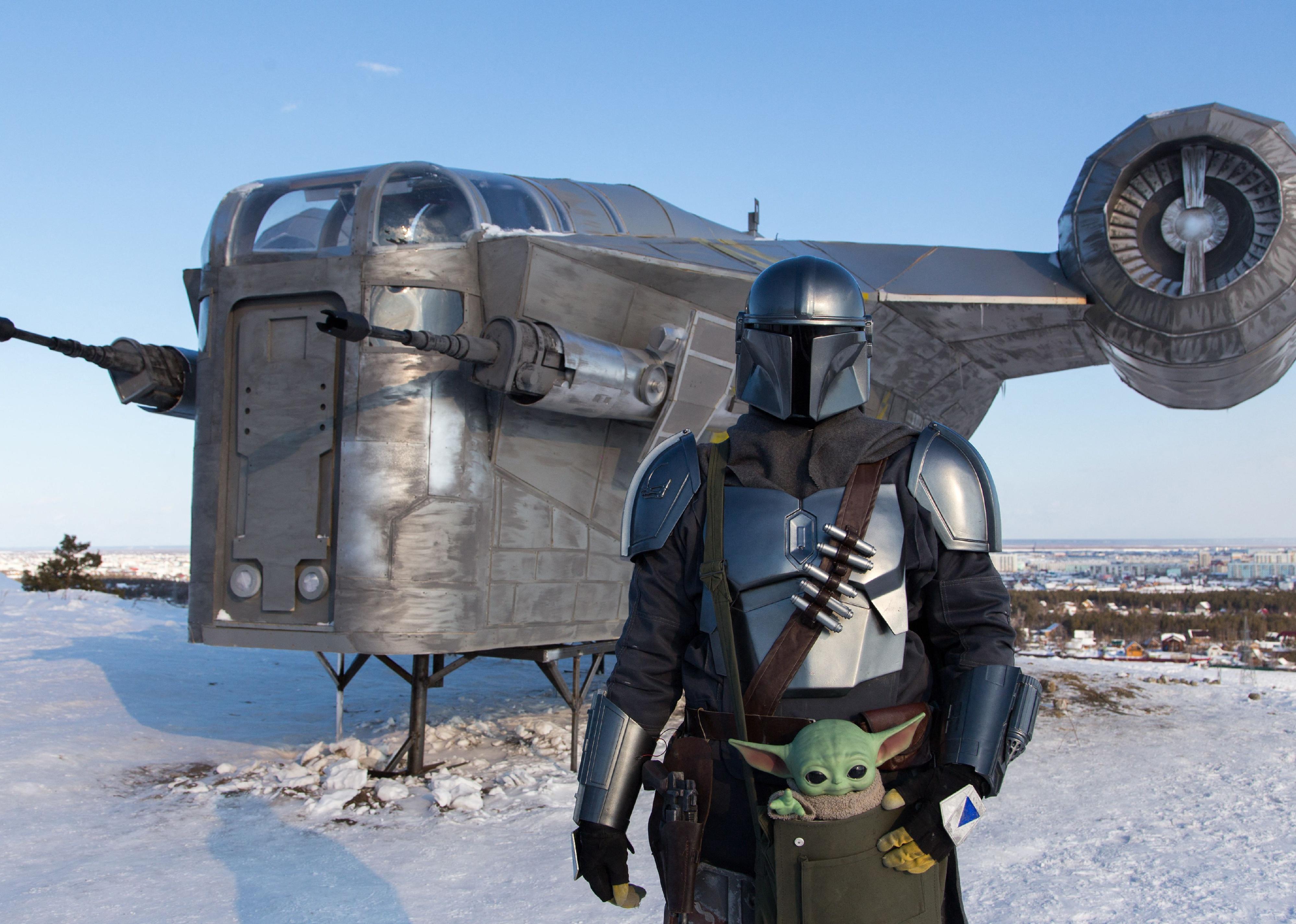 A man wearing a costume of Din Djarin poses in front of a giant replica of the Razor Crest.