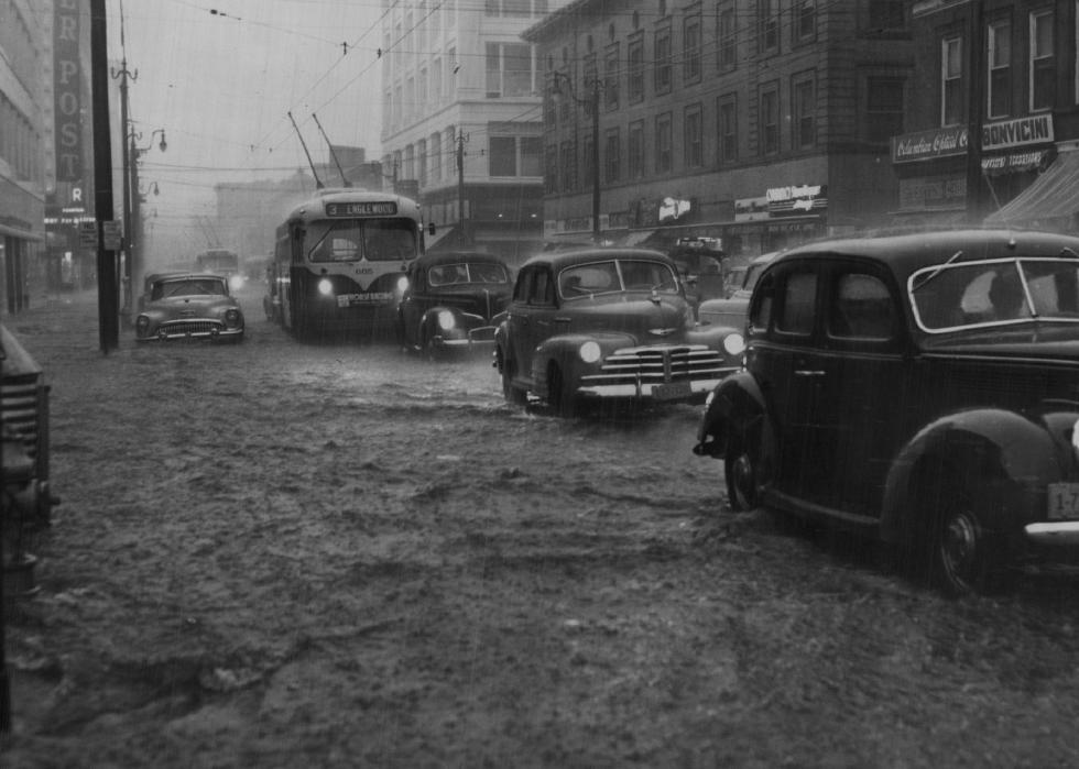 Cars and a bus drive through a flooded urban area in the rain.