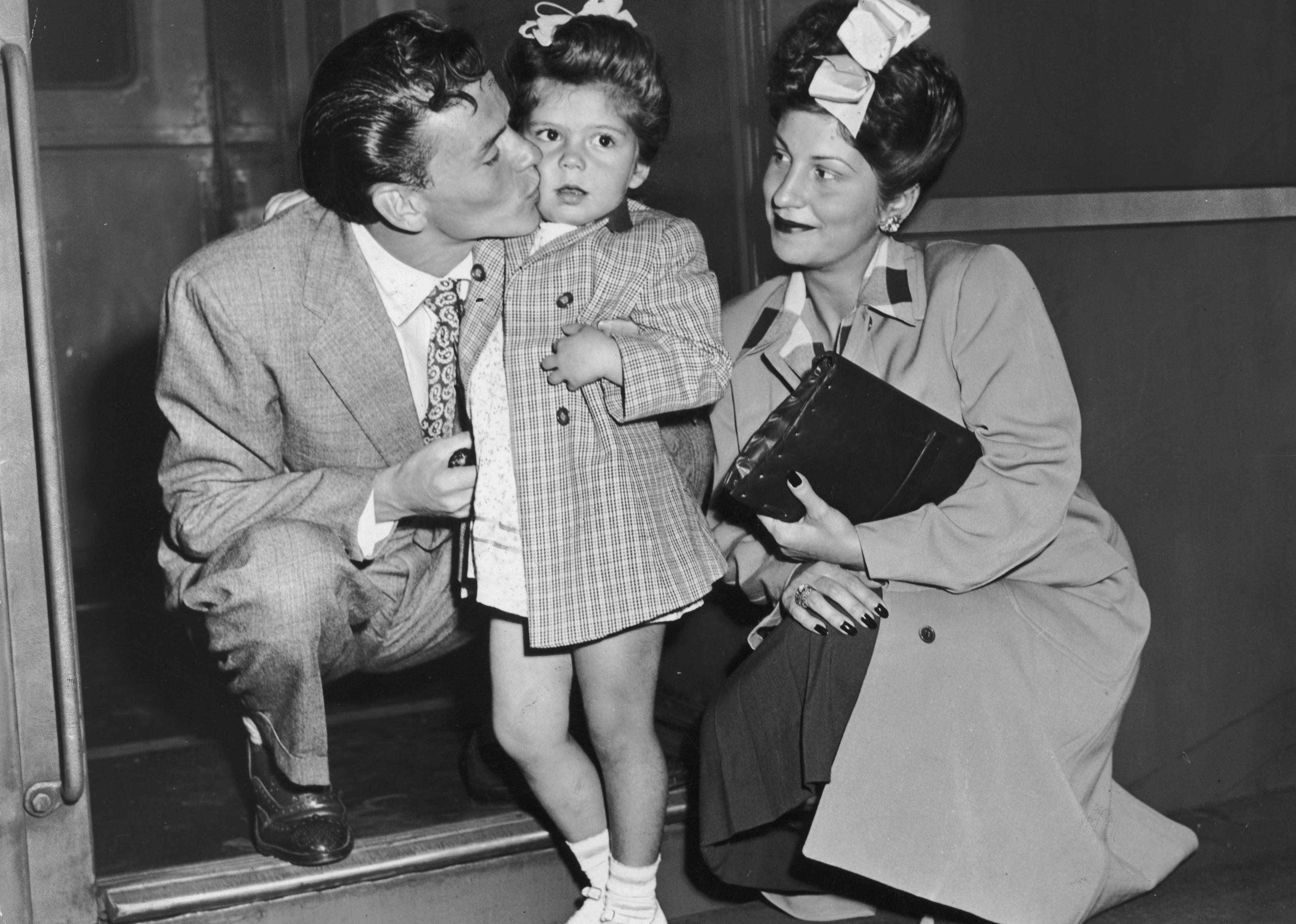 Frank Sinatra crouches to kiss his daughter, Nancy Jr. while his wife watches at a train platform. 