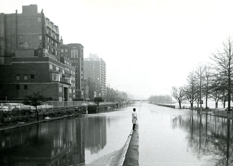 A person stands on a slightly raised bit of pavement in the middle of a flooded street.