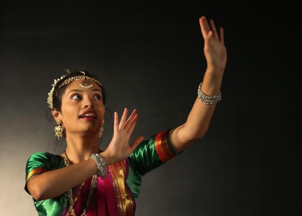 A woman wearing a brightly colored sari and jewelry dancing, looking upward with her palms lifted to the sky.