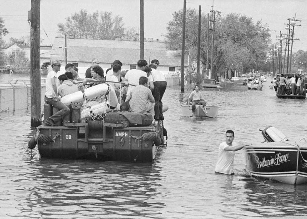 A military vehicle with at least 10 people piled on drives through waist-deep flood waters, while another two people guide their boats along the flooded street.