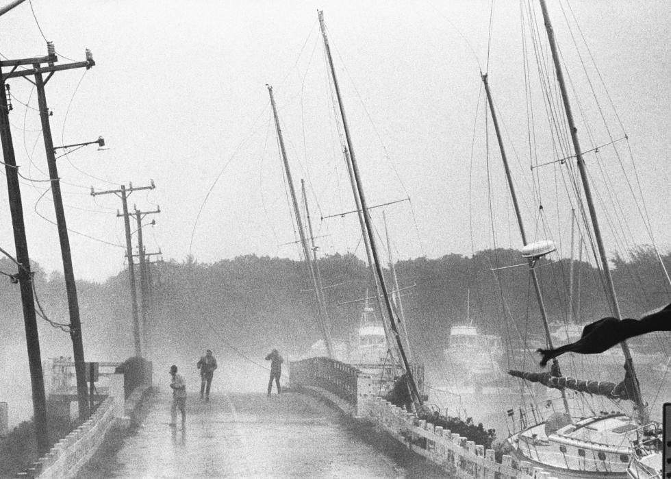 During a storm, three people walk over a bridge where several sailboats are docked.