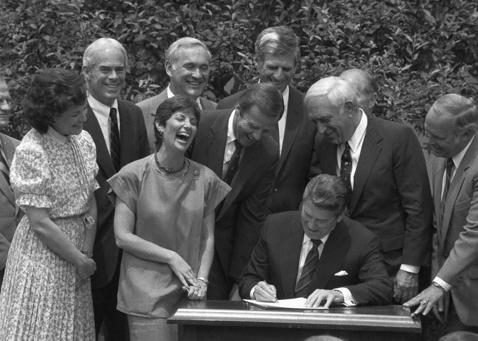 President Reagan, sat at a desk surrounded by smiling people, signs legislation raising the legal drinking age to 21 in a Rose Garden ceremony.