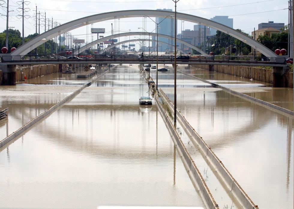 A stalled car on a mostly empty flooded highway near an urban area.