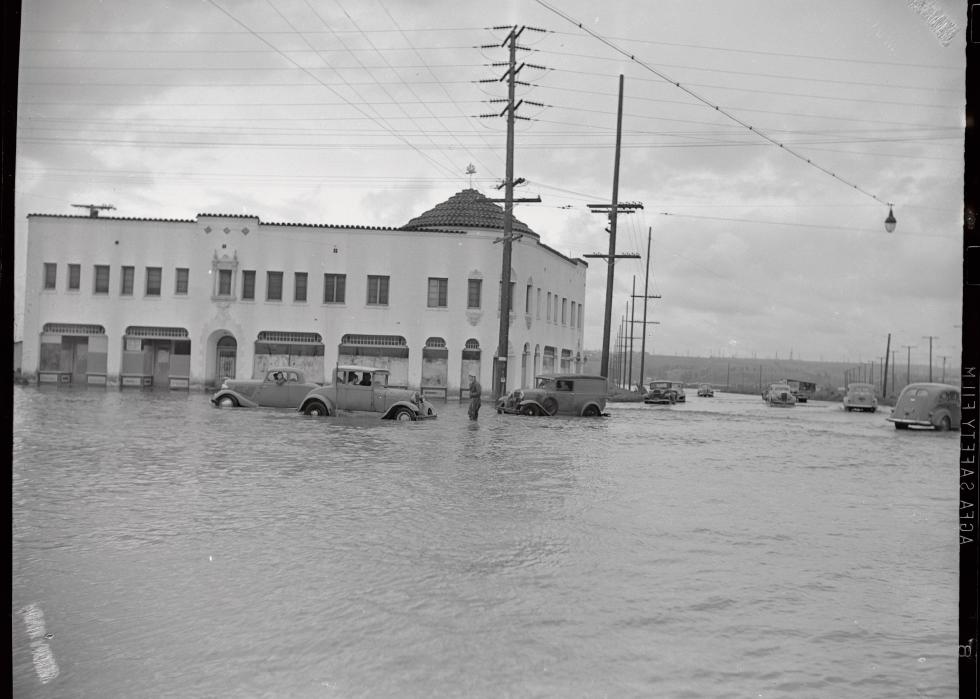 A warehouse surrounded by flood waters that come above halfway up the wheels of the cars parked.