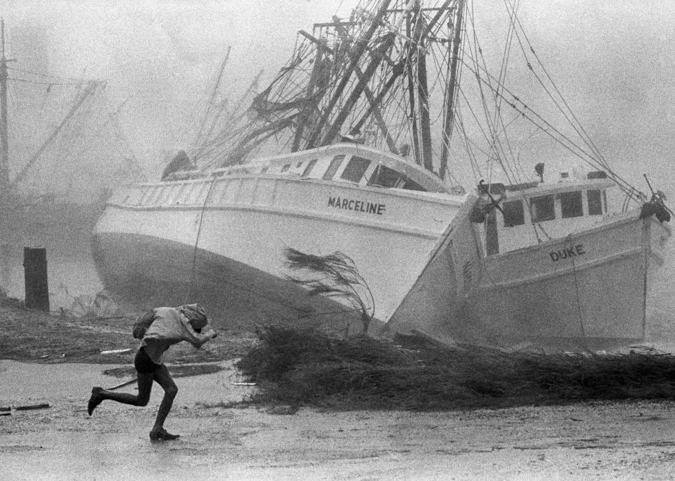 A person running through a storm next to the shore where two large fishing boats are crashed upon it.