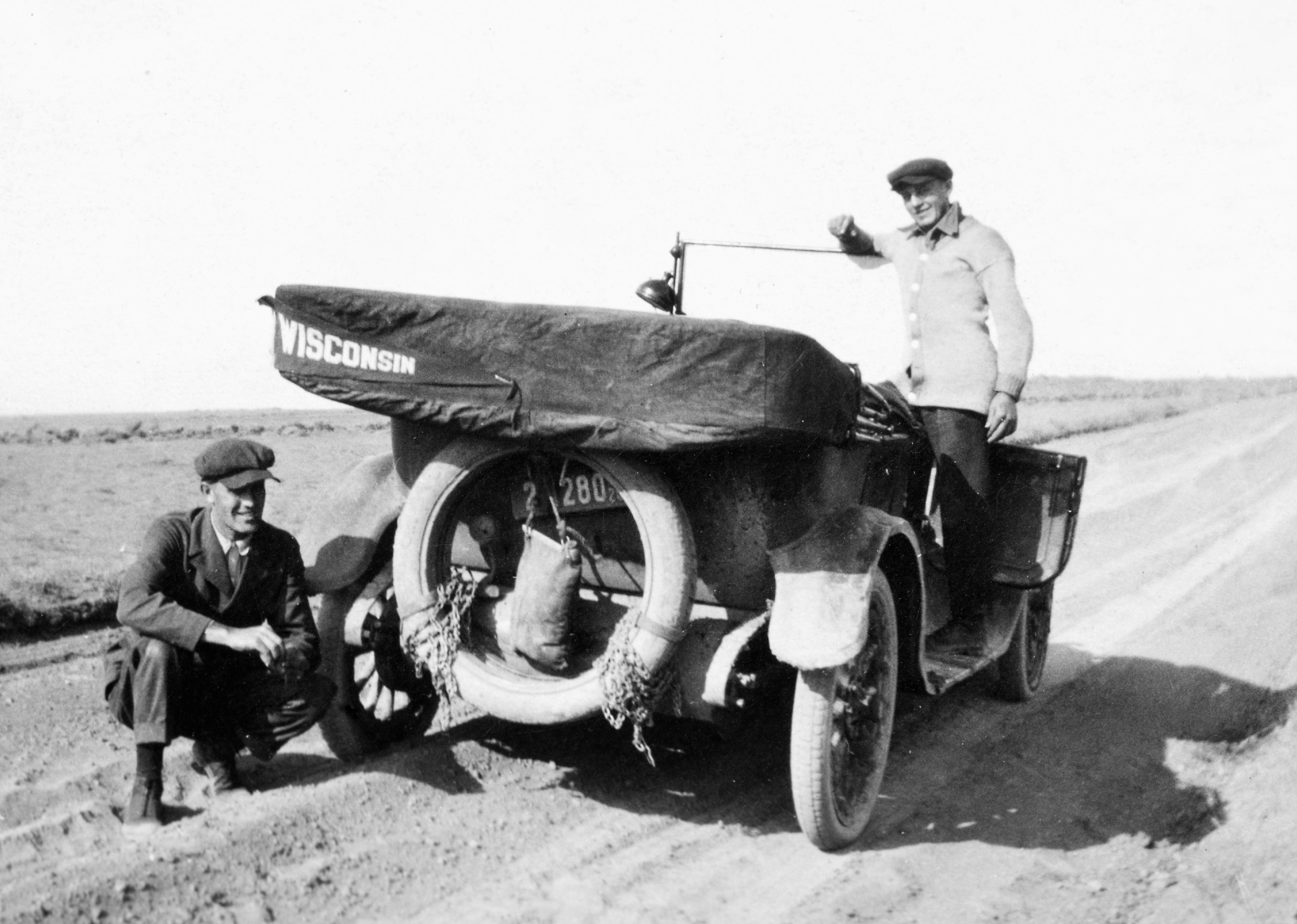 Two people on a road trip, posing next to their car with a Wisconsin flag, circa 1925.