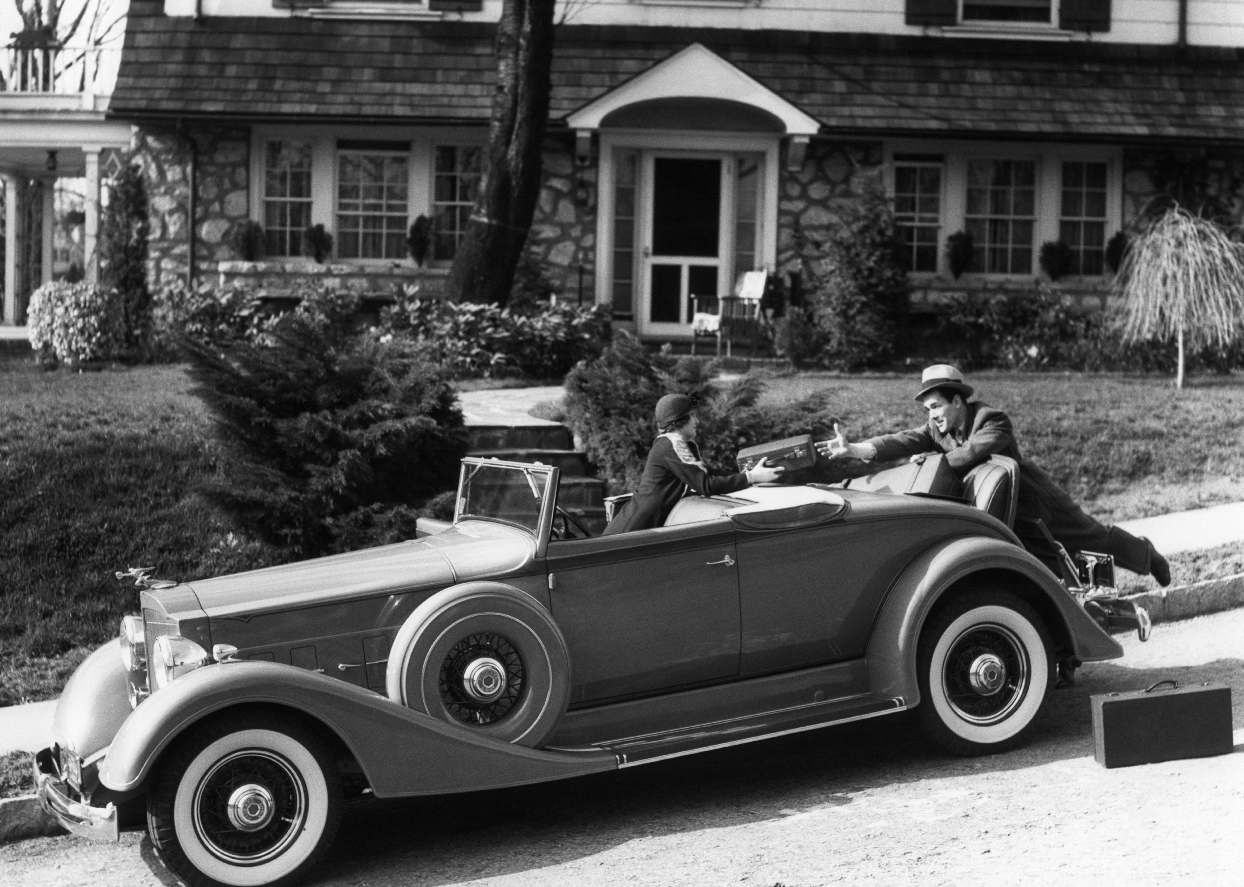 Person in car handing suitcase to someone else at the back of the car in front of a house.