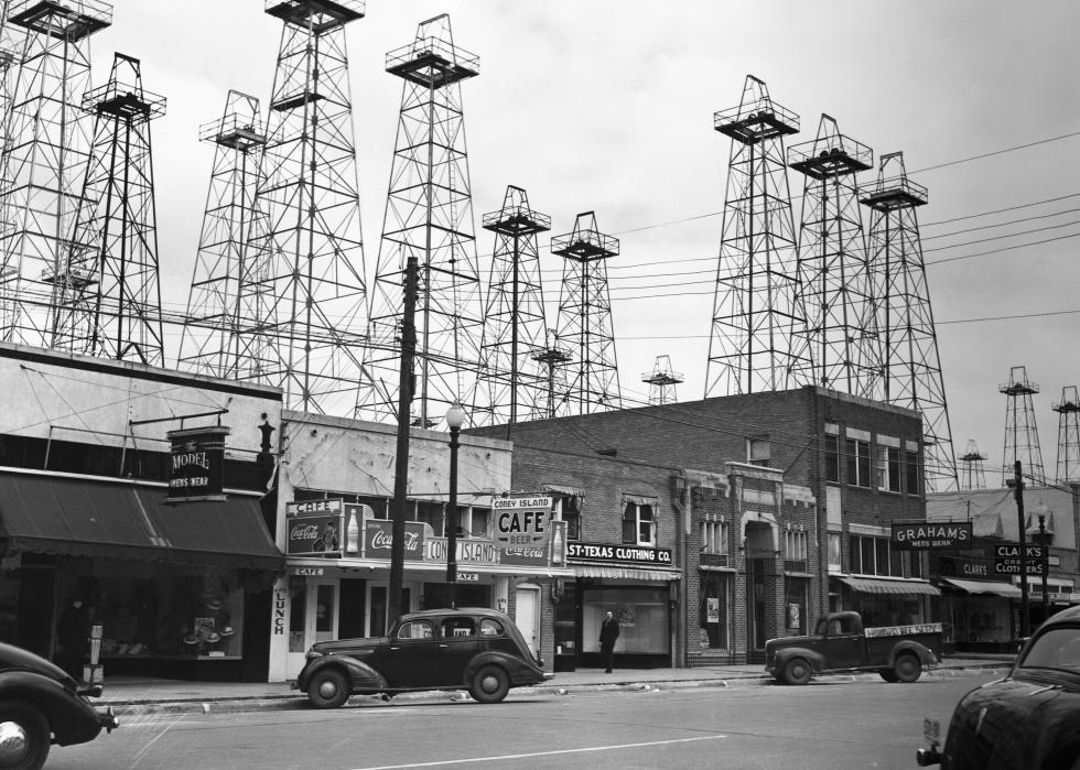 Dark clouds around a small town street with storefronts and parked cars with several transmission towers in the background. 