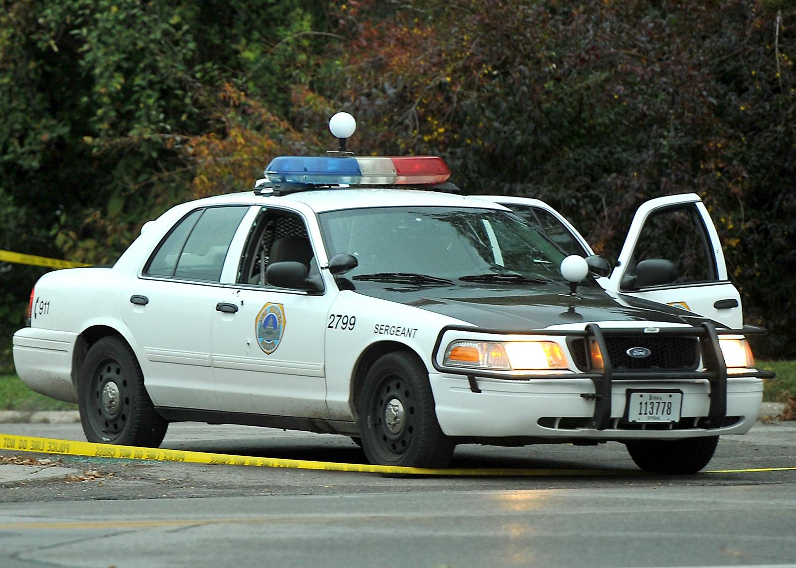 A police car with a broken window sits at a crime scene.