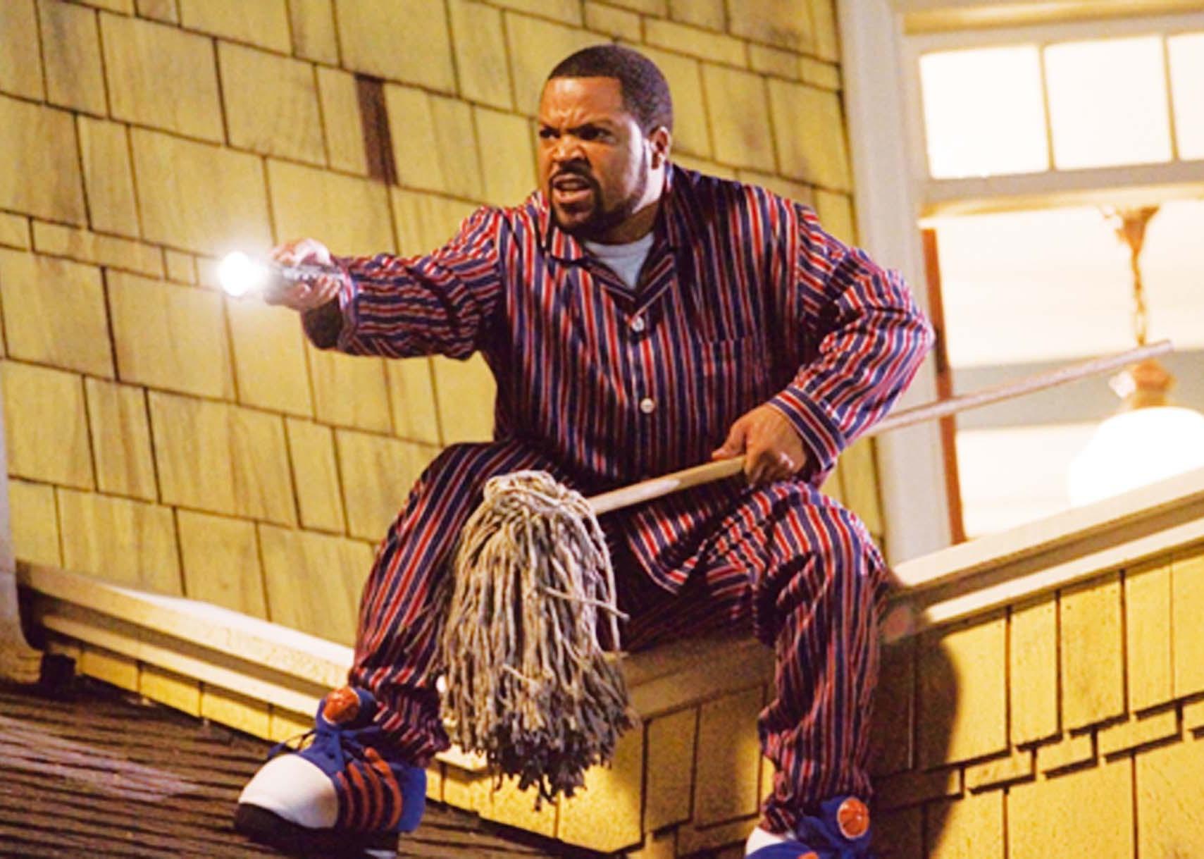 Ice Cube wearing pajamas on the roof of a house with a mop and a flashlight.