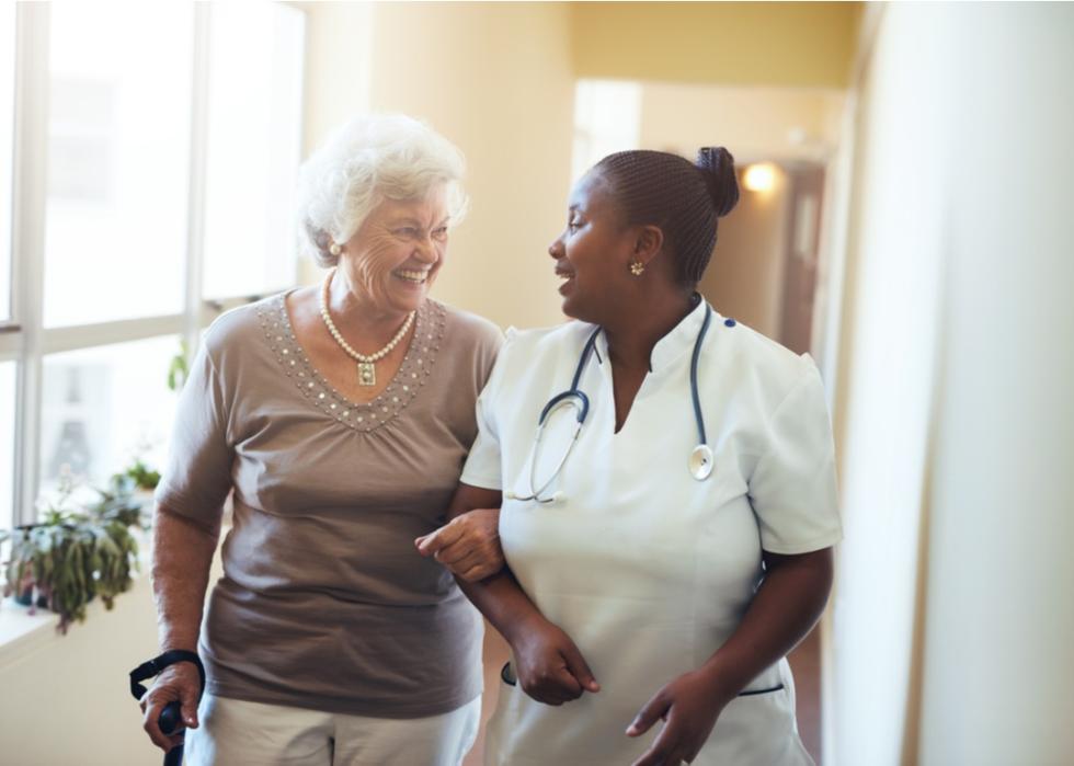 A nurse helps an elderly patient with a walker as they both smile.