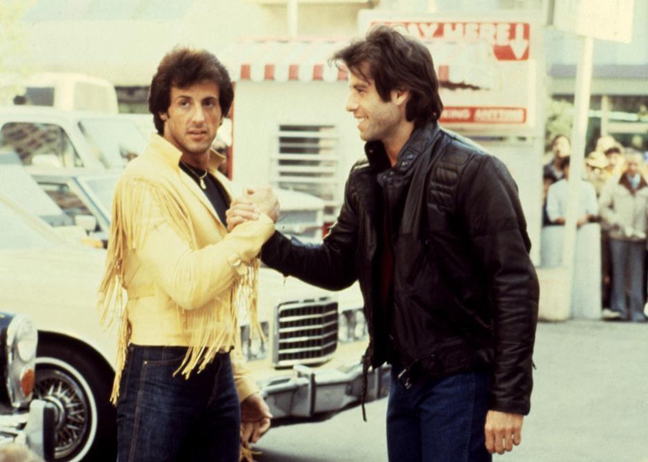 John Travolta in a black leather jacket shaking hands with Sylvester Stallone in a cream leather jacket with fringe.