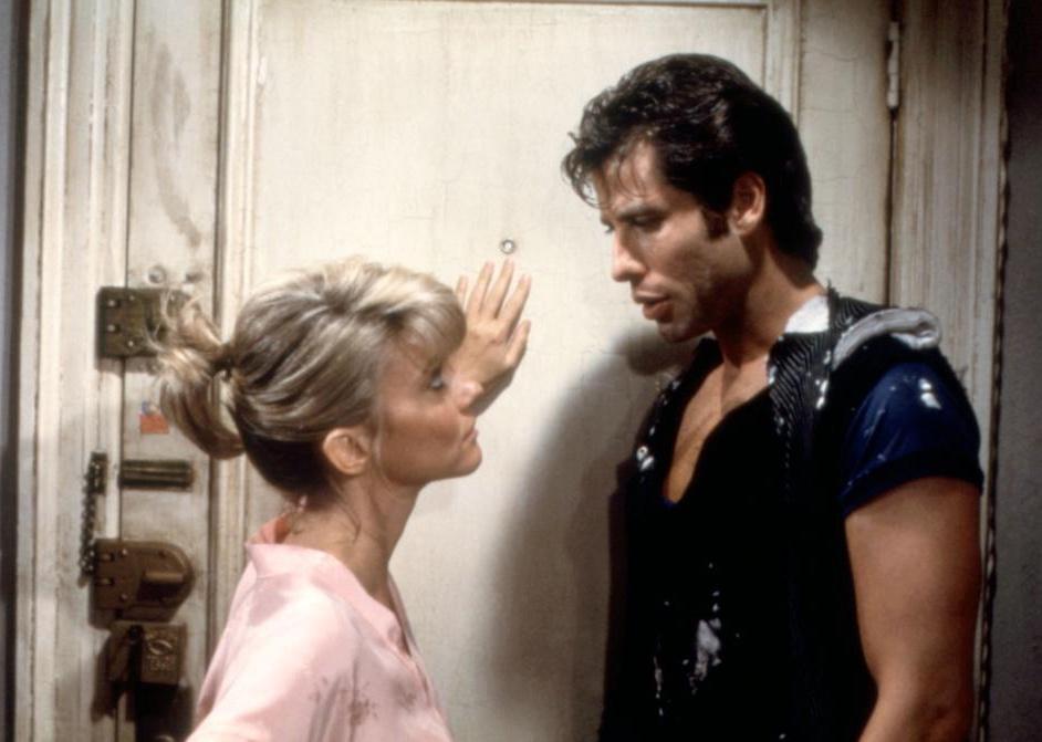 John Travolta in torn up clothes talking to a woman with short blonde hair.