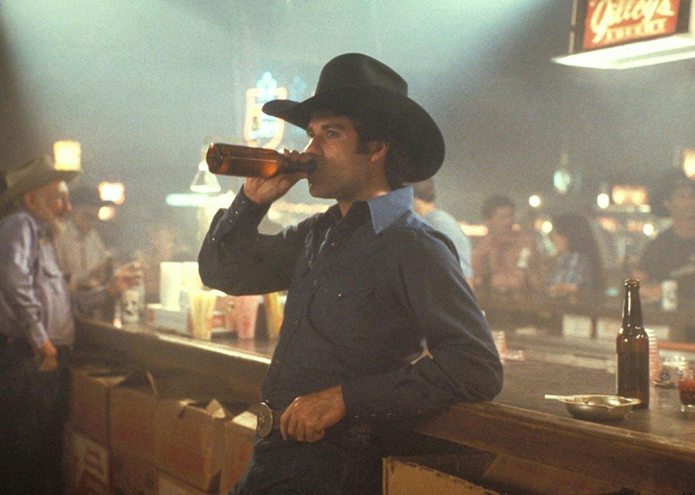 John Travolta at a bar wearing a blue western shirt and black cowboy hat.