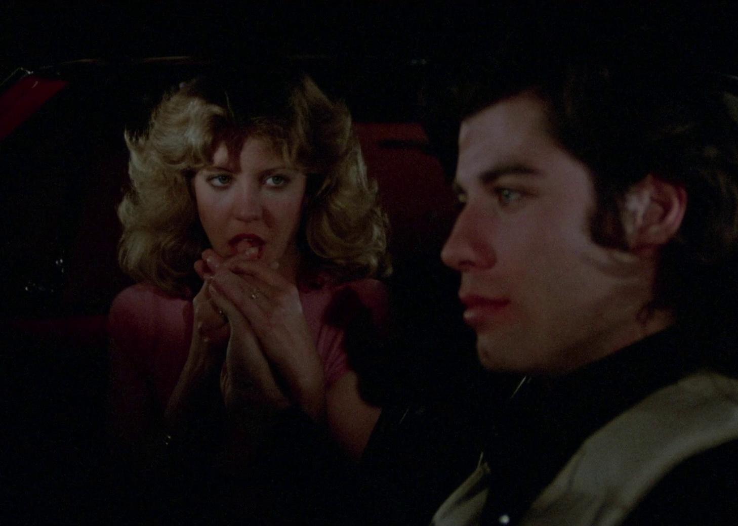 John Travolta in a car with a woman who is licking his hand.
