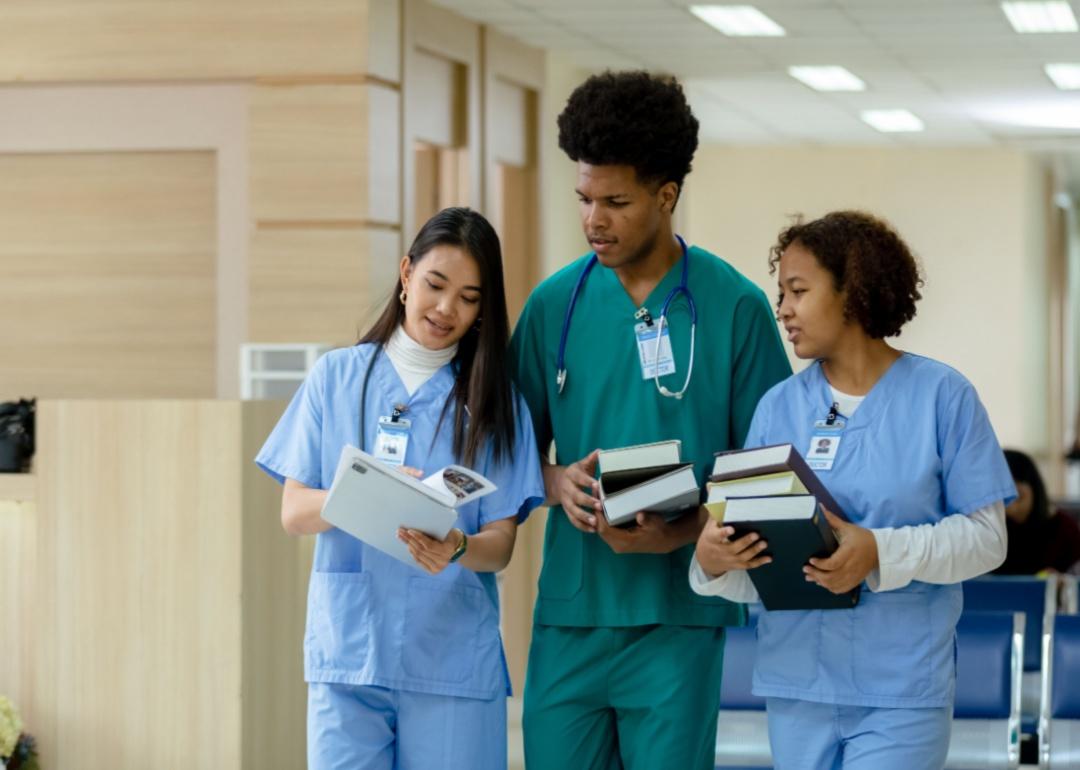 Two nursing students holding books talk with another nurse in a hospital.