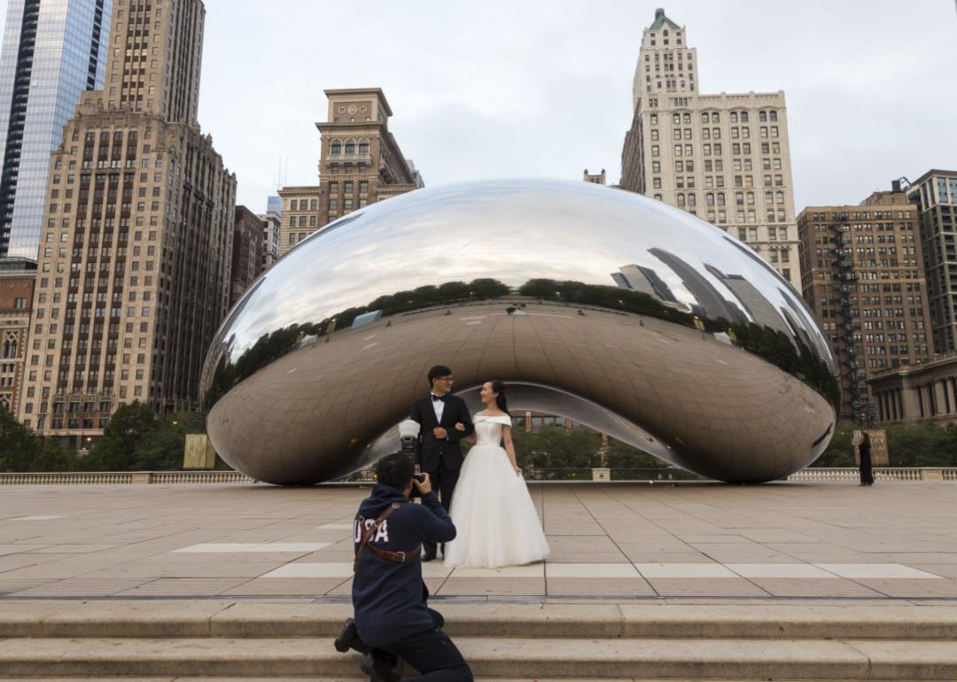 A person photographing a couple in wedding attire in front of a sculpture in downtown Chicago.