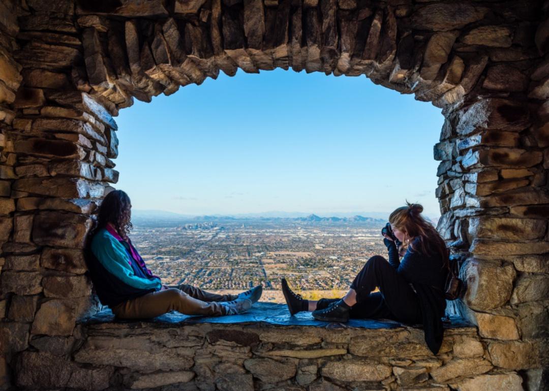 Two people sitting in a stone lookout while one of them photographs the view of Phoenix.