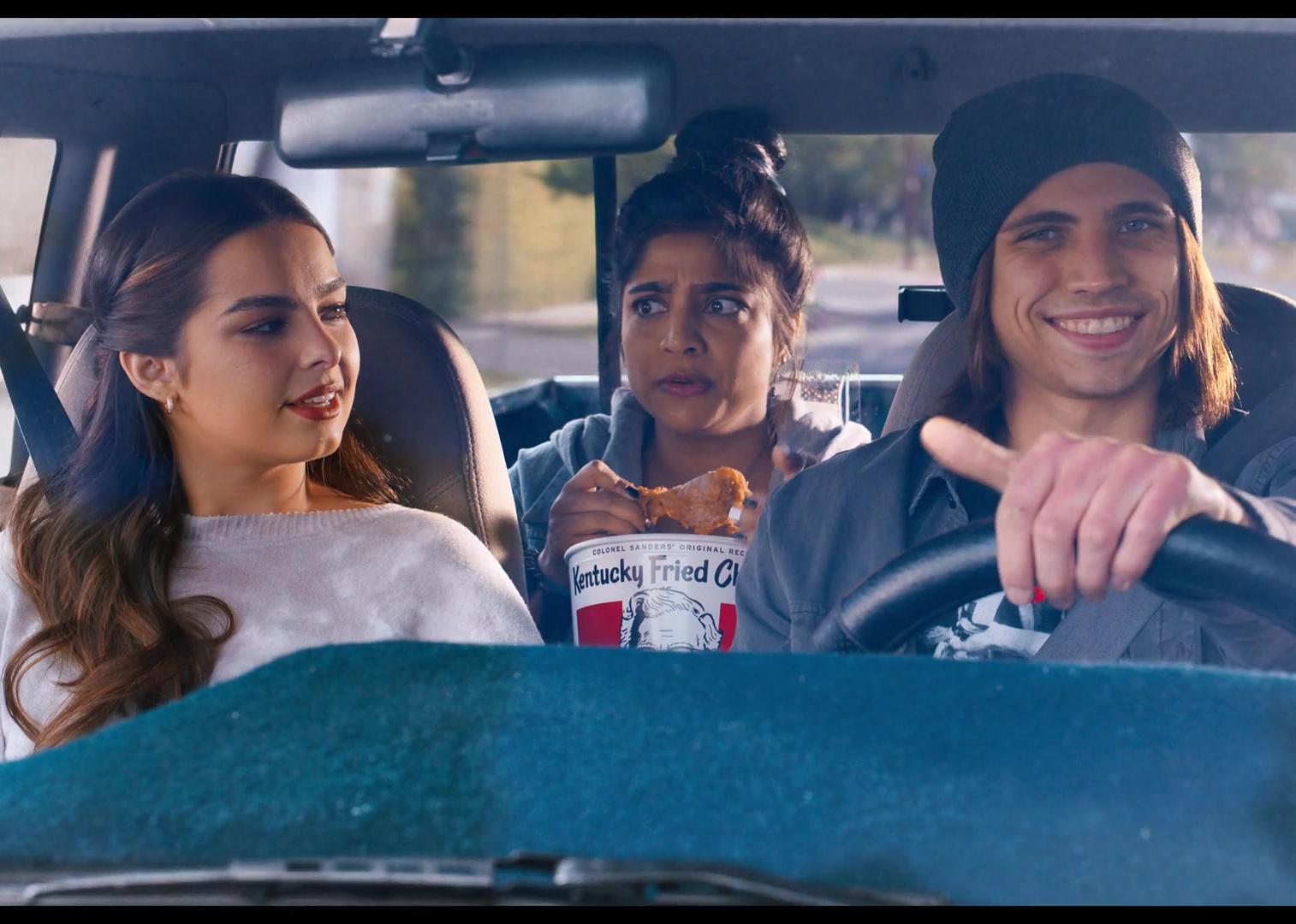 Addison Rae, in the passenger seat of a car, looking at a young man driving and smiling while a young woman in the back seat eats a bucket of fried chicken.