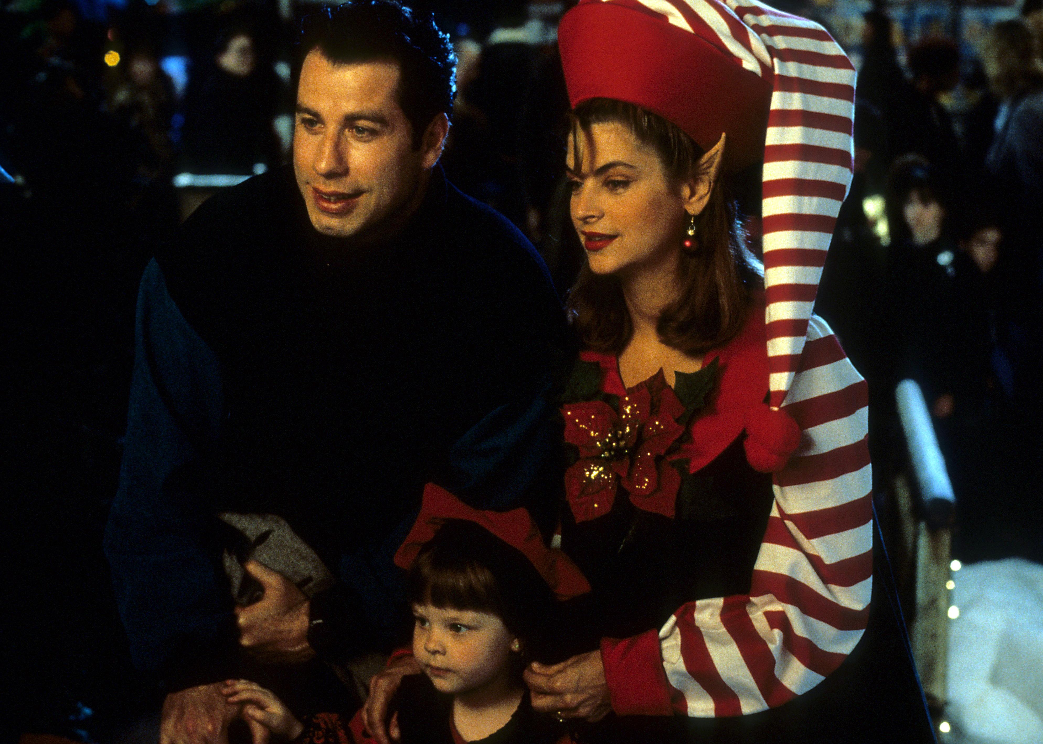 Kirstie Alley dressed as an elf taking a photo with John Travolta and a little girl.