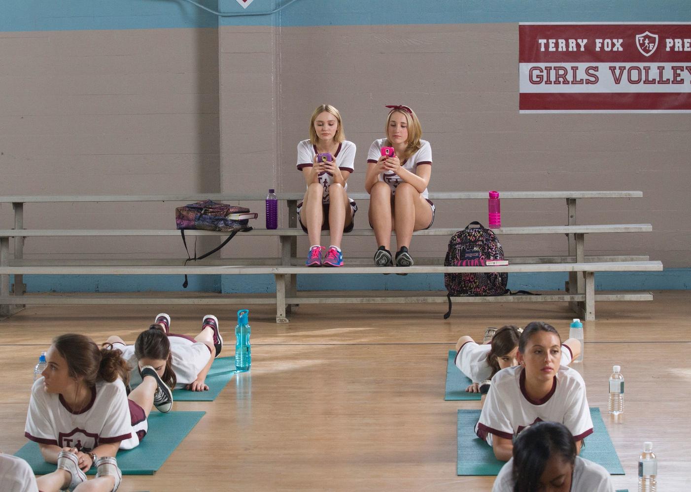 Harley Quinn Smith and Lily-Rose Depp sitting on bleachers looking at their phones while girls do yoga in a gym.