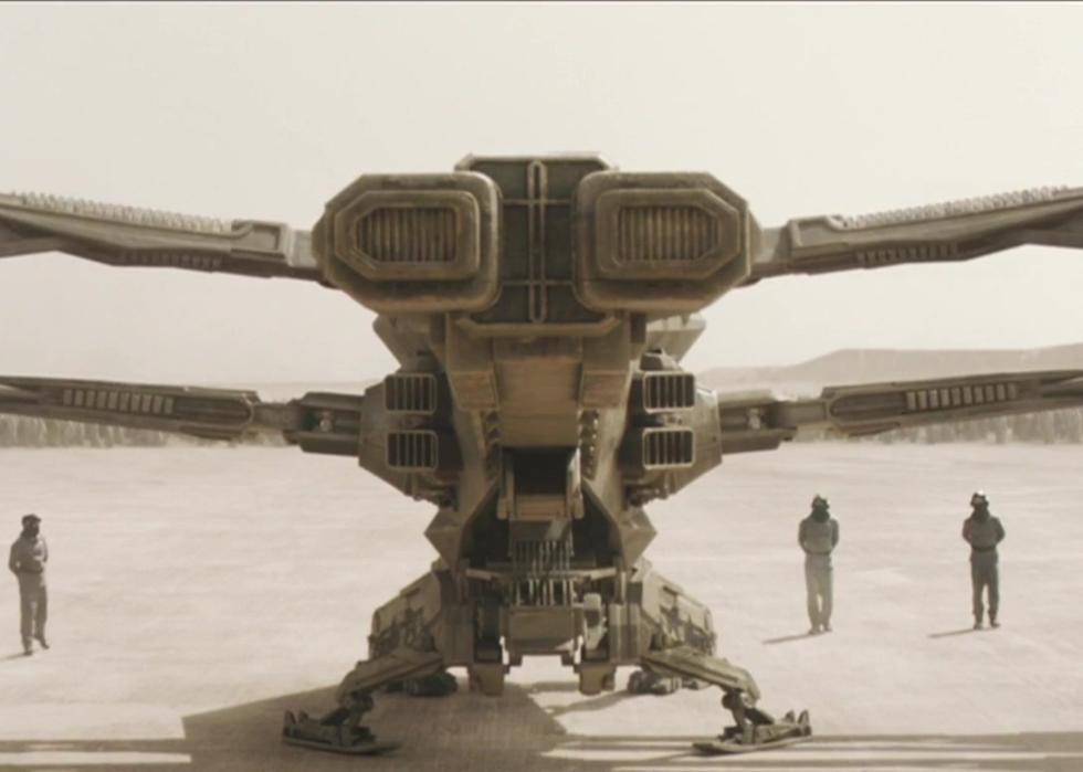 A large dragonfly shaped ship sits in the desert with an army of people in the background.