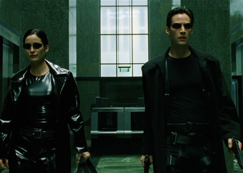 Man and woman dressed in all black with suglasses and guns walk through a corporate building.
