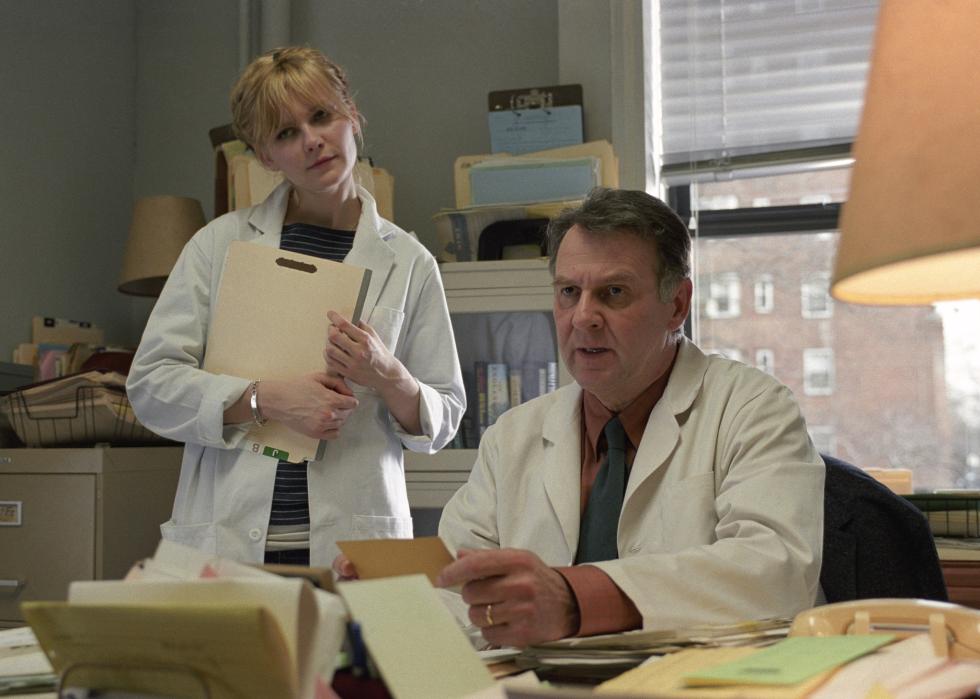 An older man and a young woman wearing white lab coats in an office.