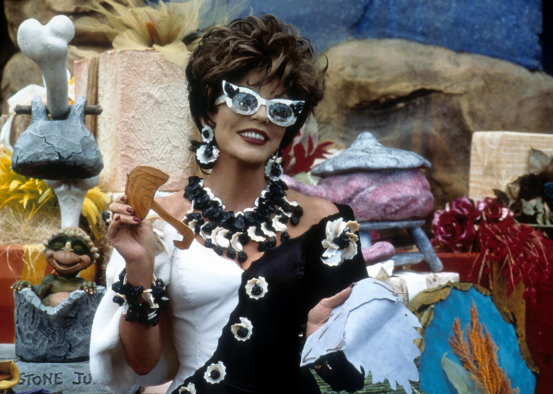 Joan Collins in a black and white dress and big white glasses smiling.