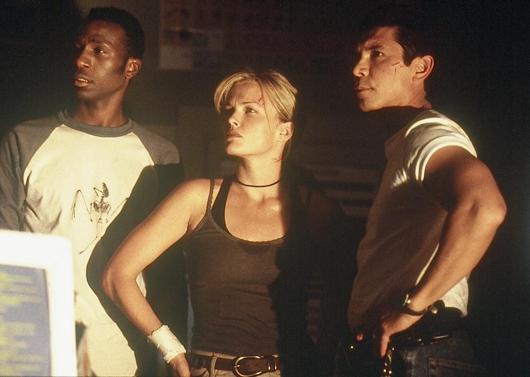 Dina Meyer, Lou Diamond Phillips, and Leon stand looking inquisitively at something.