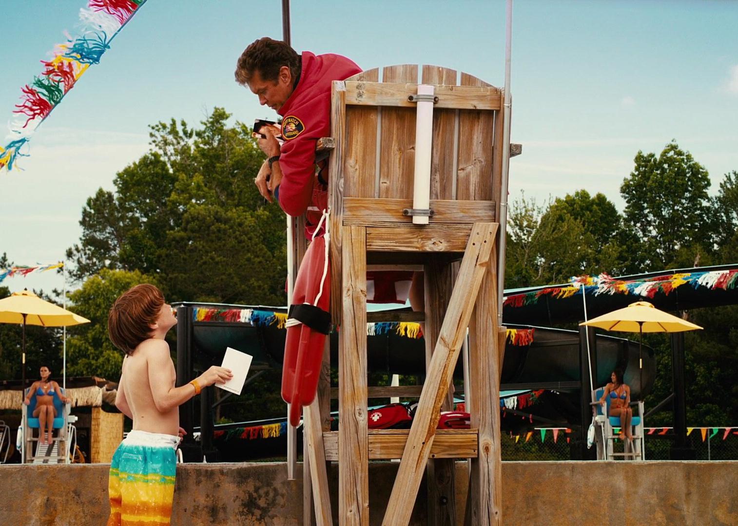 David Hasselhoff in a lifegaurd stand talking to a little boy with two women in bikinis in the background.