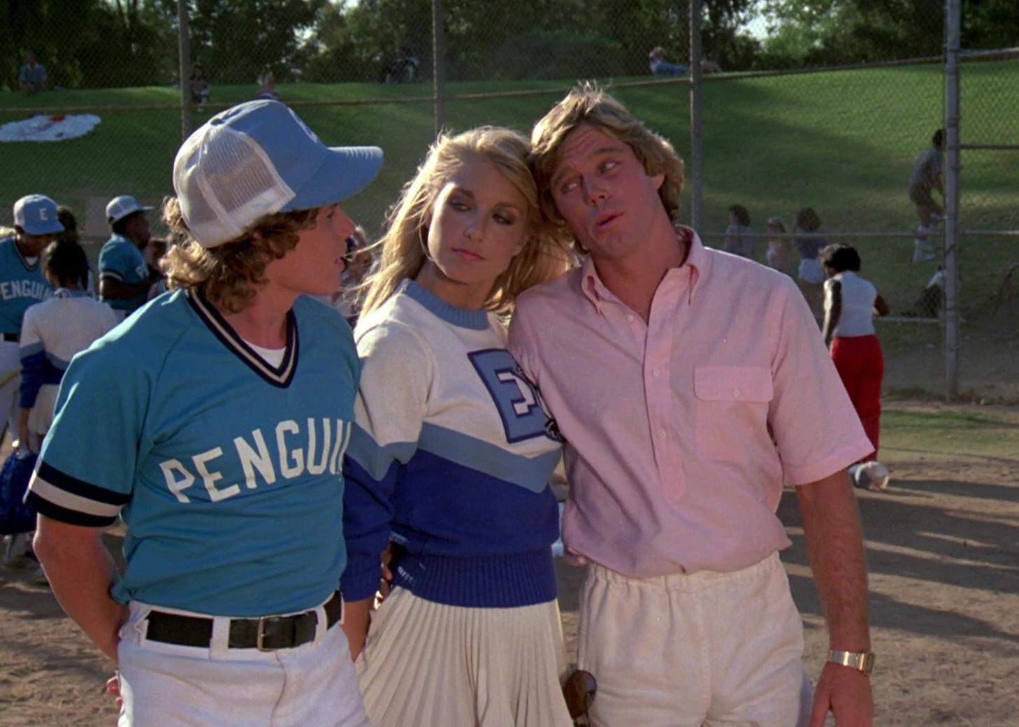A blonde cheerleader in between a baseball player and a blonde guy in a pink button down shirt on the baseball field.