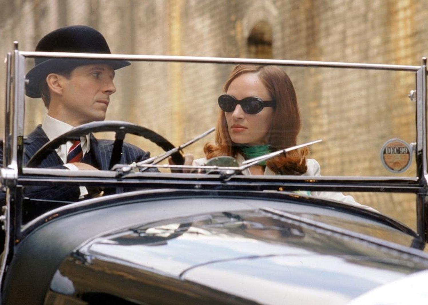 Ralph Fiennes in a pinstripe suit and top hat and Uma Thurman in cat eye glasses driving in a classic car.