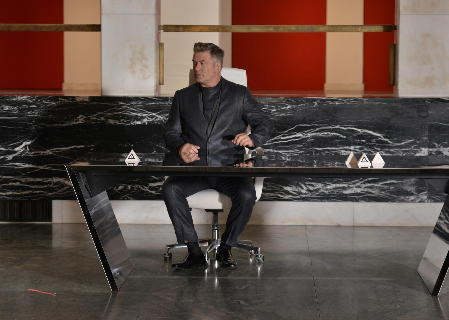 Alec Baldwin in a black suit sitting at a modern desk in front of a black and white marble wall.