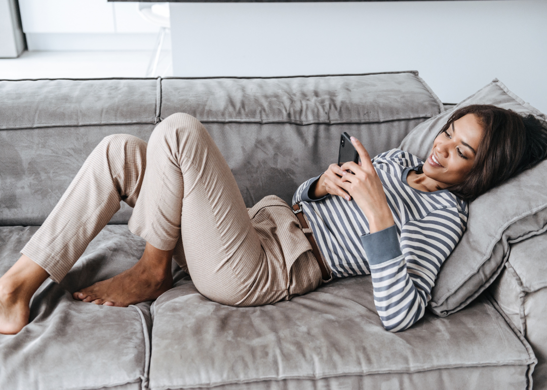 A woman lying on a gray velvet couch smiling and looking at her phone.