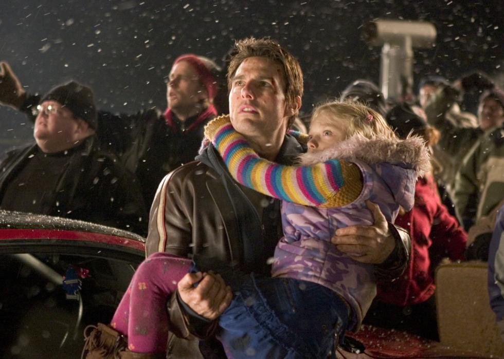 Tom Cruise holds a little girl during a storm in a crowd of people all looking up at something.