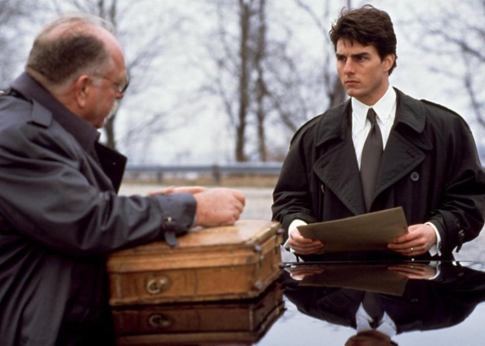 Two men wearing dark coats over suits and looking at files outside a car.