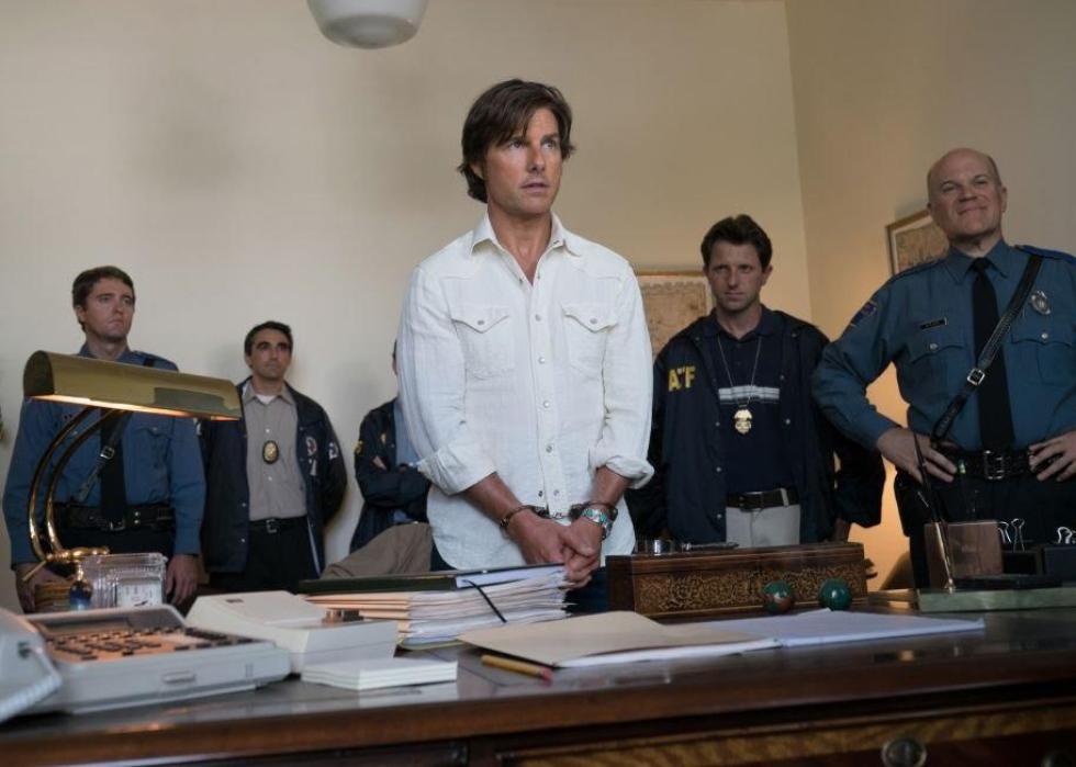 Tom Cruise stands in handcuffs in an office in front of a line of police officers.