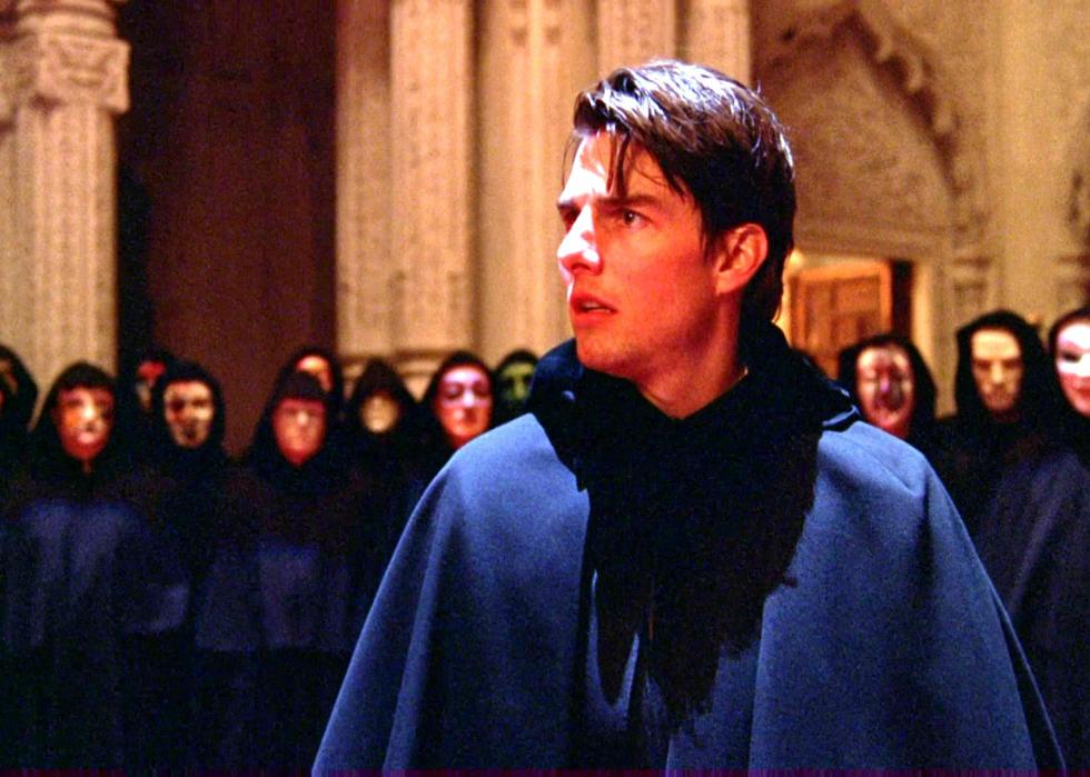 Tom Cruise looks surprised in a room surrounded by people wearing black robes and masks.
