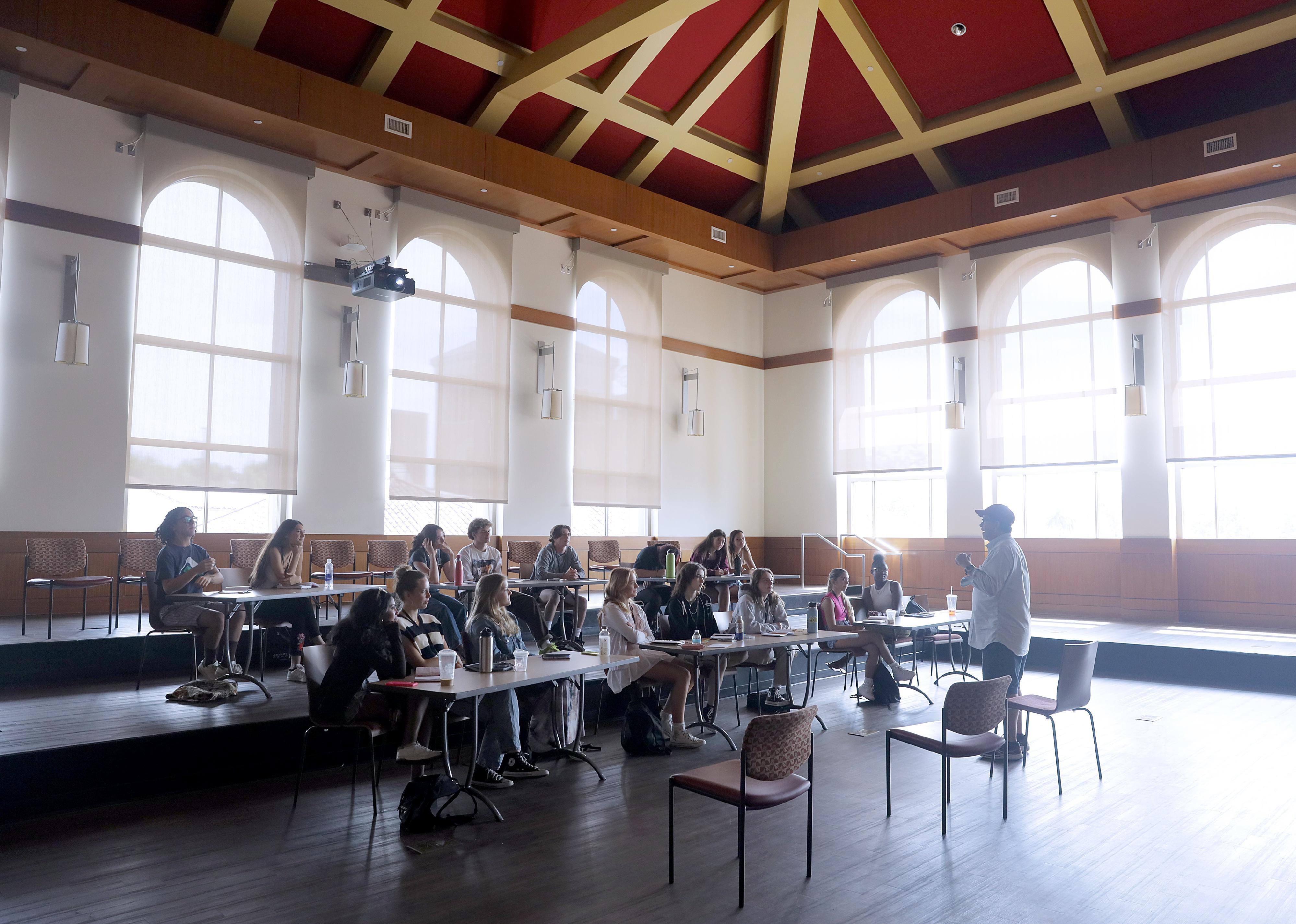 Students listen to a tutor in a large open room lined with windows.