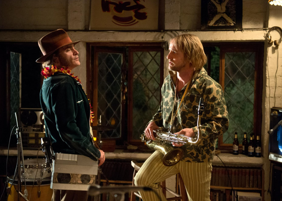 Joaquin Phoenix, wearing a recording device on a strap, standing next to Owen Wilson holding a saxophone in Inherent Vice.