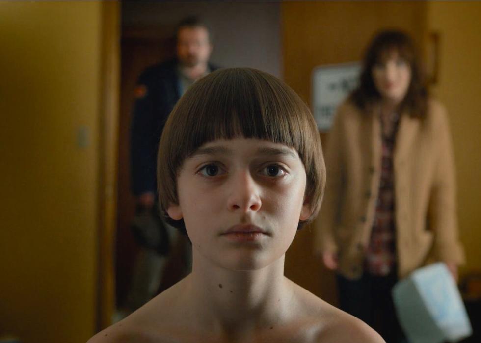 Noah Schnapp sitting staring forward with Winona Ryder and David Harbour in the background.