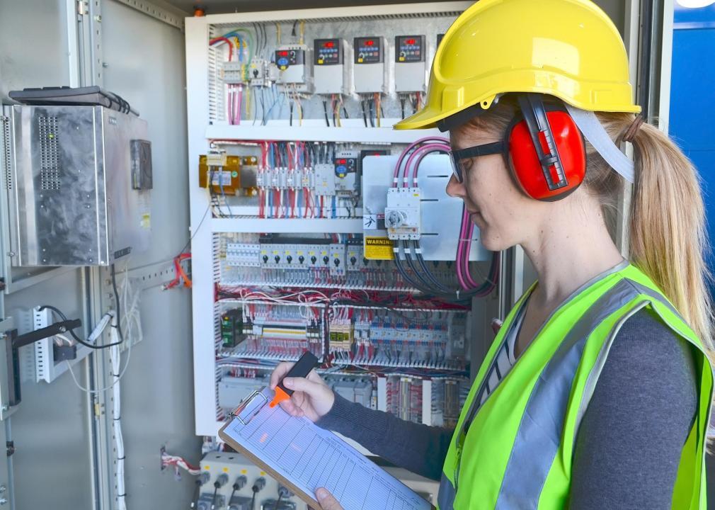 Woman electrical engineer wearing safety gear while writing on a clipboard.