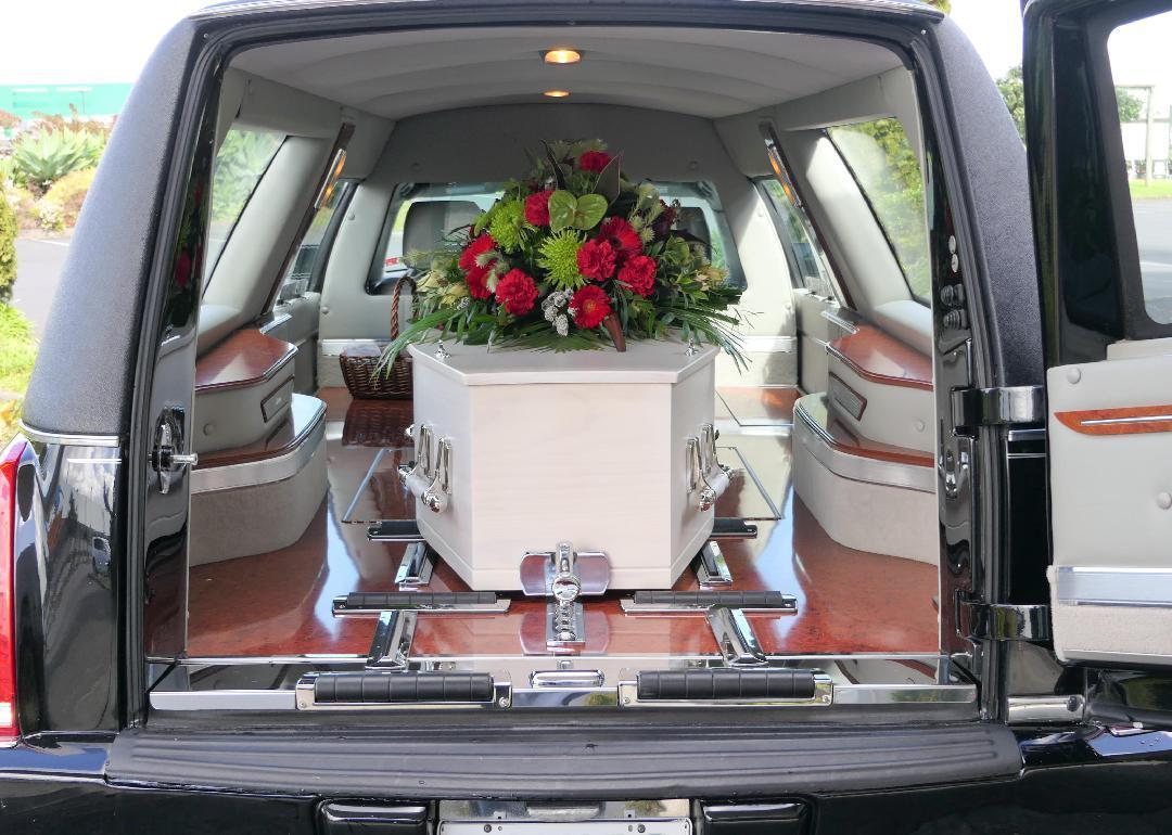 A white casket with a floral arrangement on top, in the back of a hearse. 