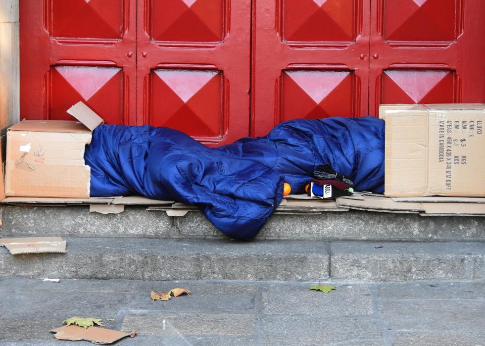Two people sleeping, with their heads partially covered by cardboard boxes, on steps in front of red doors.